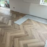 Jasna podłoga winylowa w jodełkę z częściowo ułożonym szarym płytkiem ceramicznym na krawędzi