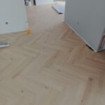 Jasna podłoga winylowa w wzorze jodełki z widocznymi narzędziami i materiałami budowlanymi w tle