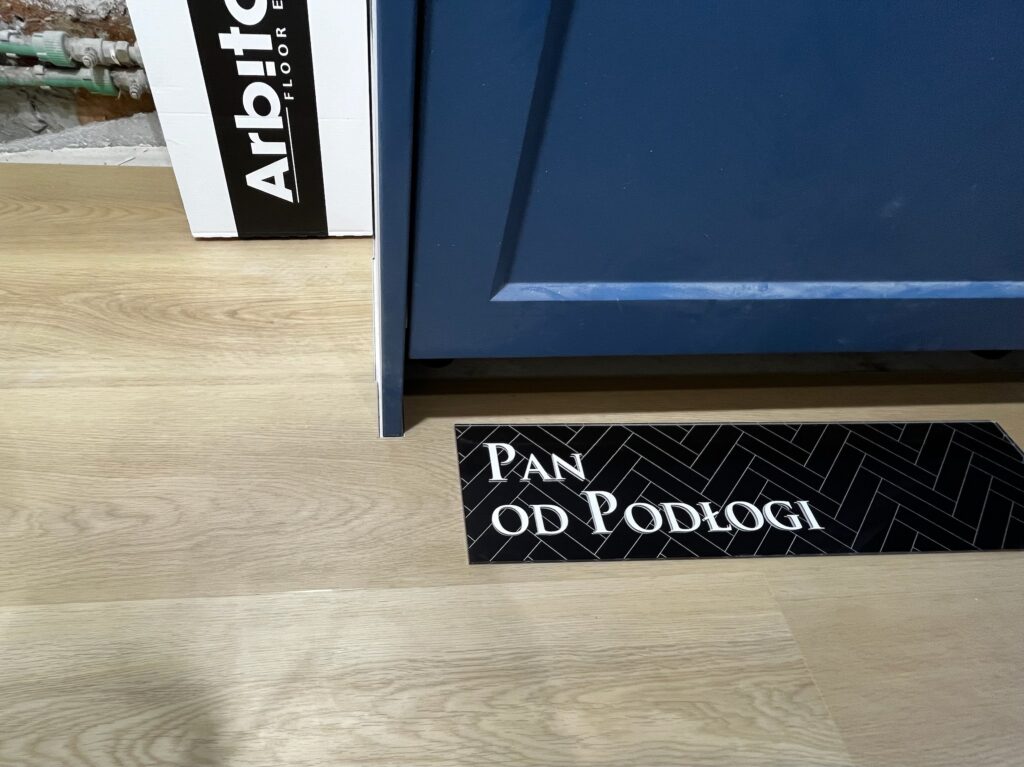 Czarny panel podłogowy z napisem "PAN OD PODLOGI" obok niebieskiej zabudowy i kartonu "Arbiton.floor".