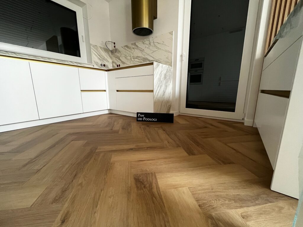 Drewniana podłoga w jodełkę z czarnym panelem z napisem "PAN OD PODLOGI" przy ścianie.